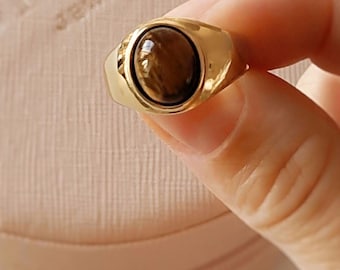 May include: A gold-colored ring with a brown, oval-shaped stone. The ring has a smooth, polished finish and is held in a person's hand. The ring is a piece of jewelry.