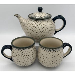 May include: A ceramic teapot and two matching mugs featuring a cream-colored base with a dense pattern of small dark blue dots. The teapot has a rounded body, a curved spout, a lid with a knob, and a dark blue handle. Each mug has a rounded shape.
