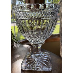 May include: A clear glass pedestal bowl featuring intricate diamond-cut patterns around the upper rim and base. The bowl has a flared opening, vertical fluted detailing along the body, and a square-shaped foot with a radial starburst design.