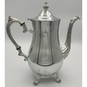 May include: A polished silver-tone metal coffee pot featuring a tall, tapered body, a curved handle with two black circular accents, a long ornate spout, a removable lid with a finial top, and three decorative feet at the base of the vessel.
