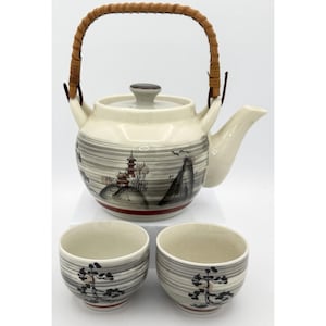 May include: A ceramic teapot with a woven brown handle and a matching lid, featuring a gray landscape illustration with a pagoda and mountains. Two small matching ceramic cups are included, each decorated with a black tree motif.