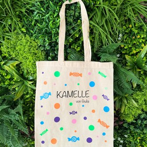 Puede incluir: Una bolsa de lona beige con un dise&ntilde;o de caramelos coloridos. La bolsa tiene un asa blanca y el texto "KAMELLLE von Giulia" impreso en la parte delantera.