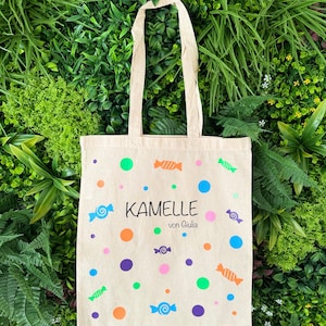 Puede incluir: Una bolsa de lona beige con un dise&ntilde;o de caramelos coloridos. La bolsa tiene un asa blanca y el texto "Kamelle von Giulia" impreso en la parte delantera.