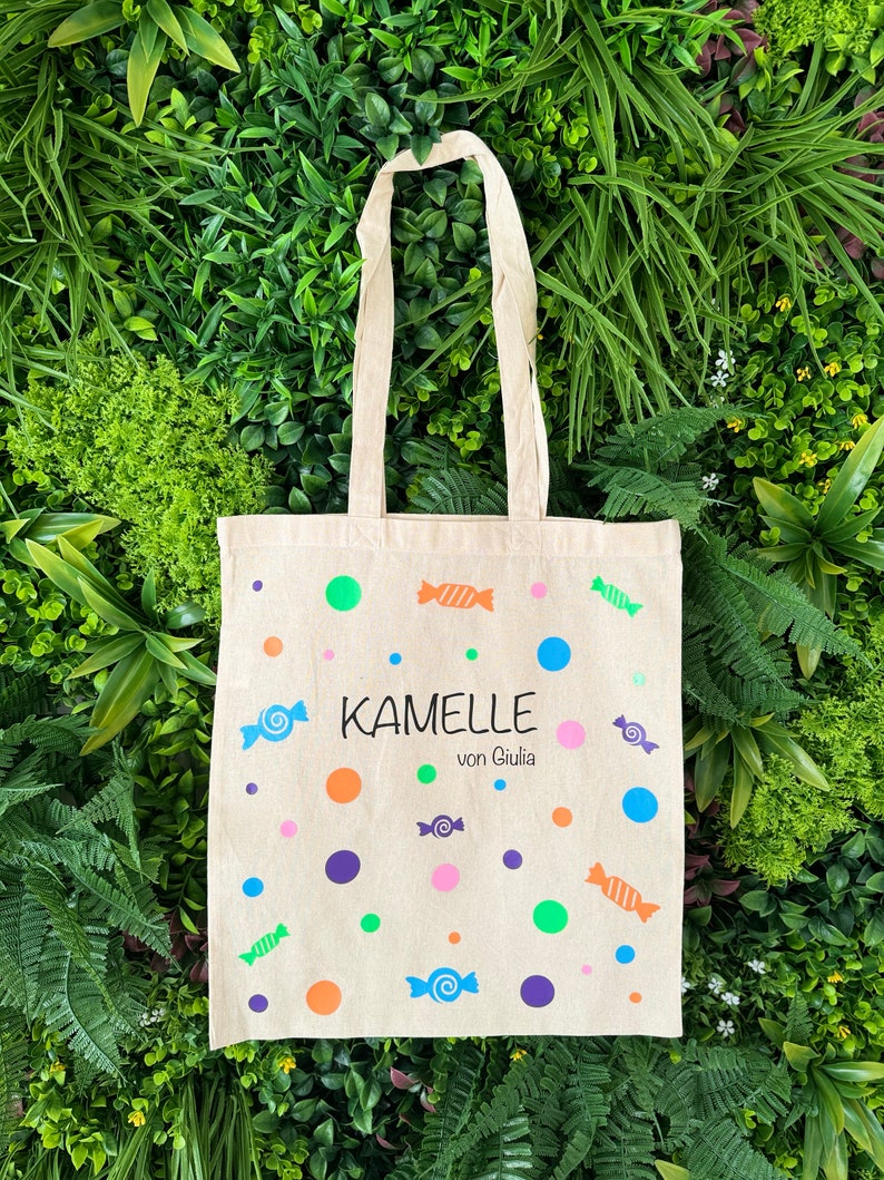 Puede incluir: Una bolsa de lona beige con un dise&ntilde;o de caramelos coloridos. La bolsa tiene un asa blanca y el texto "Kamelle von Giulia" impreso en la parte delantera.