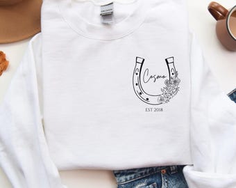May include: A white long-sleeve sweatshirt with a horseshoe design, the name "Cosmo" in script, and the text "EST 2018". The sweatshirt is paired with blue jeans, a brown hat, and a cup of coffee.
