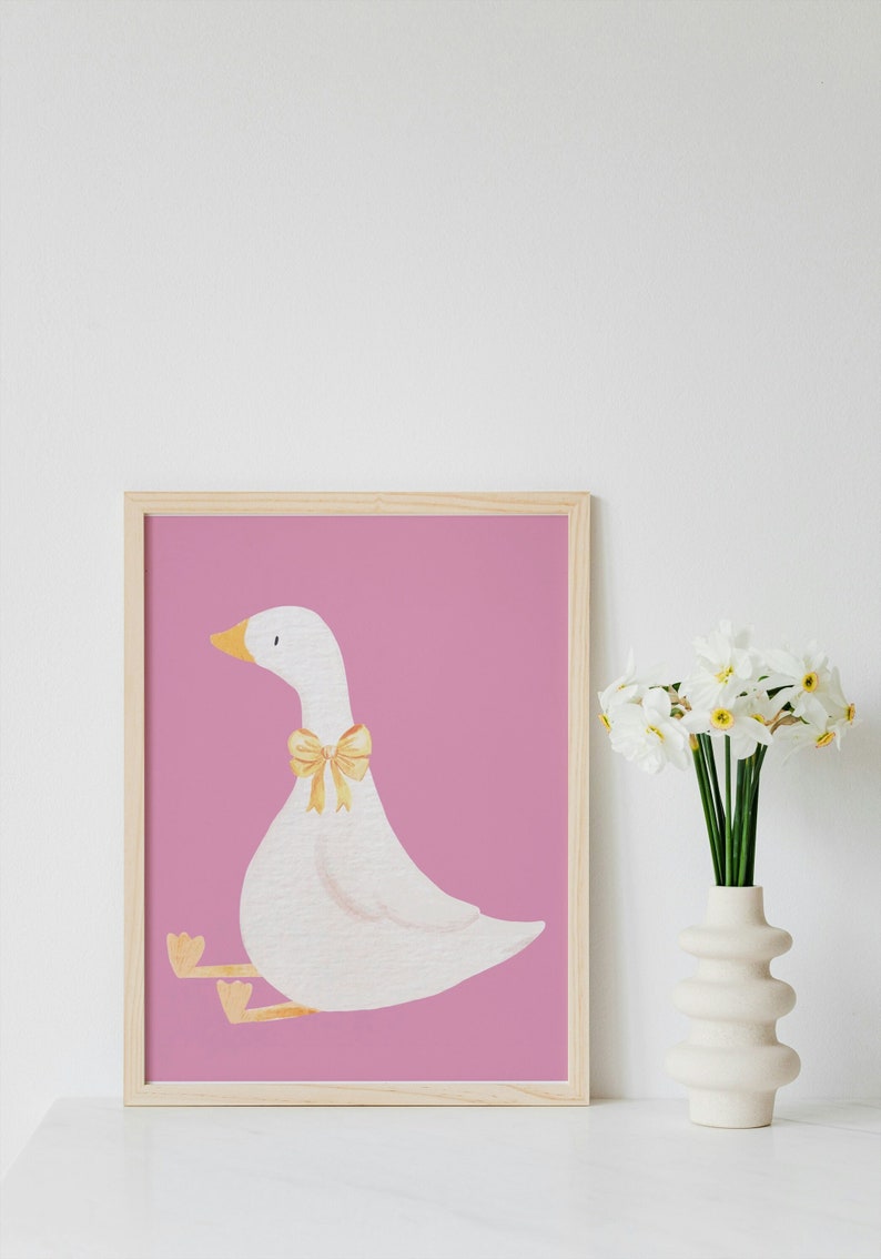 May include: A framed print of a white goose with a yellow bow sitting on a pink background. The print is in a light wood frame and is sitting on a white surface.