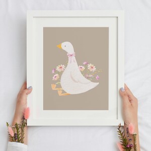 May include: A white framed print with a light brown background featuring a white goose with a pink bow sitting on a bed of pink and yellow flowers.