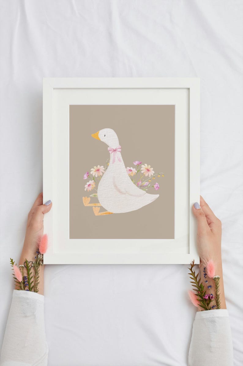 May include: A white framed print with a light brown background featuring a white goose with a pink bow sitting on a bed of pink and yellow flowers.