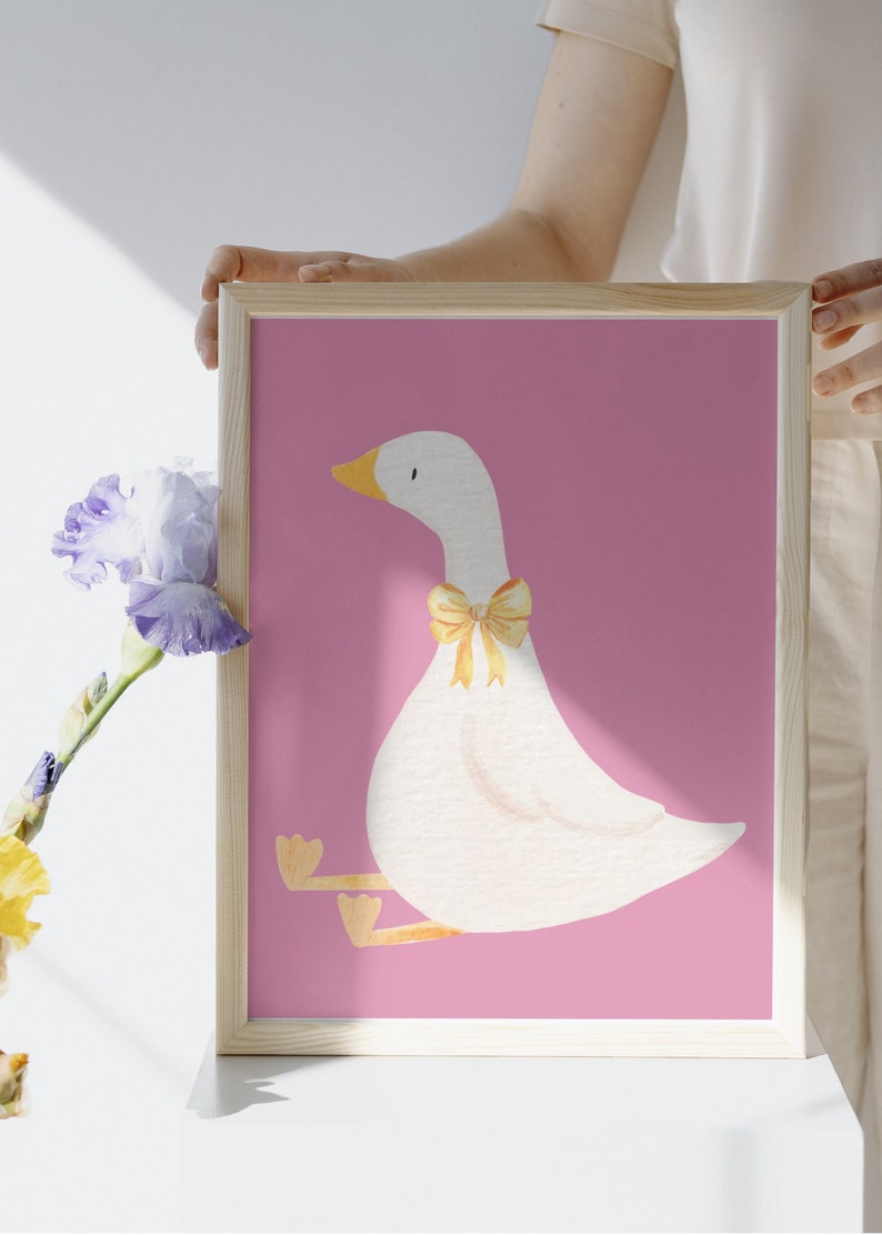 May include: A framed print of a white goose with a yellow bow sitting on a pink background.