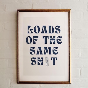 Può includere: Stampa incorniciata con il testo blu navy "LOADS OF THE SAME SHIT" in un carattere stilizzato. È inclusa una piccola illustrazione di una molletta per il bucato. La cornice è dorata, su un muro di mattoni bianchi.
