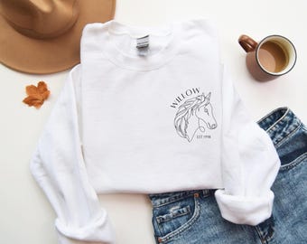 May include: A white crewneck sweatshirt with a black line drawing of a horse's head and the word "WILLOW" above it. The sweatshirt also has the text "EST 1998" below the horse graphic. A brown hat, a leaf, a mug of coffee, and blue jeans are also in the image.