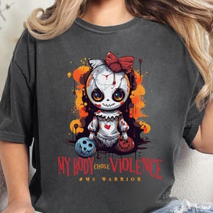 May include: Dark gray t-shirt featuring a cartoon doll with a red bow and heart. The doll is surrounded by orange and yellow splatters. Text on the shirt reads "MY BODY CHOSE VIOLENCE" and "#MS WARRIOR".