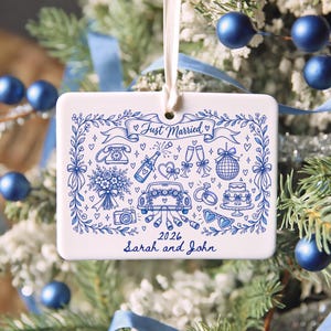 May include: A rectangular white ceramic ornament hanging from a cream ribbon, featuring blue line art of wedding icons including a car, cake, rings, and champagne. The text reads Just Married, 2026, and Sarah and John in blue script.