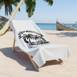 May include: White beach towel with a black graphic of palm trees and the text "Hey Moonin'" on a wooden beach chair. The chair is on a sandy beach with a boat and ocean in the background.