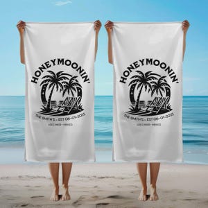 Two people standing on a sandy beach, each holding a matching white beach towel. The towels feature a black tropical graphic with palm trees, stars, and two lounge chairs under the word “Honeymoonin’” in bold text. Below the image, the towels are personalized with the text “The Smith’s – EST 06-01-2025” and “Los Cabos – Mexico.” The ocean and blue sky create a serene vacation backdrop.