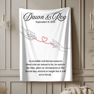May include: White throw blanket with black text that reads "Dawn & Roy" and "September 11, 2021". The blanket features a black line drawing of two hands connected by a red thread with a heart at the center. The text below the drawing reads "An invisible red thread connects those who are meant to be, no matter the time, place or circumstances. The thread may stretch or tangle but it will never break."