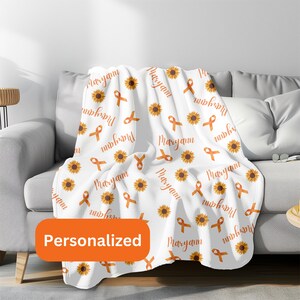 May include: White fleece throw blanket with a repeating pattern of orange ribbons and yellow sunflowers. The blanket is personalized with the name "Maryann" printed throughout the design. The blanket is displayed folded on a couch.