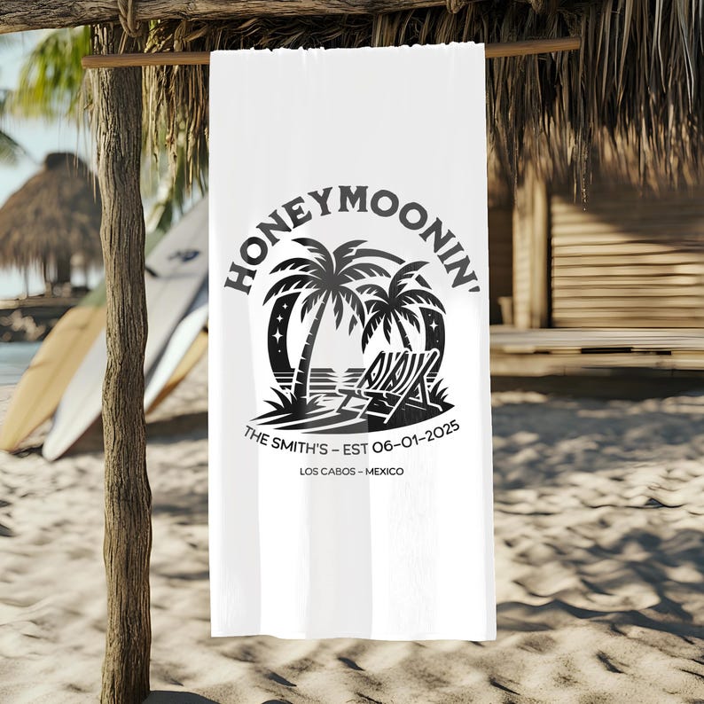 A white beach towel hanging from a wooden tiki-style cabana on a sandy beach. The towel displays a black tropical design with palm trees, two lounge chairs, and stars under the word “Honeymoonin’.” Below the graphic, the towel is personalized with “The Smith’s – EST 06-01-2025” and “Los Cabos – Mexico.” Surfboards lean against the cabana in the background, completing the laid-back tropical vibe.