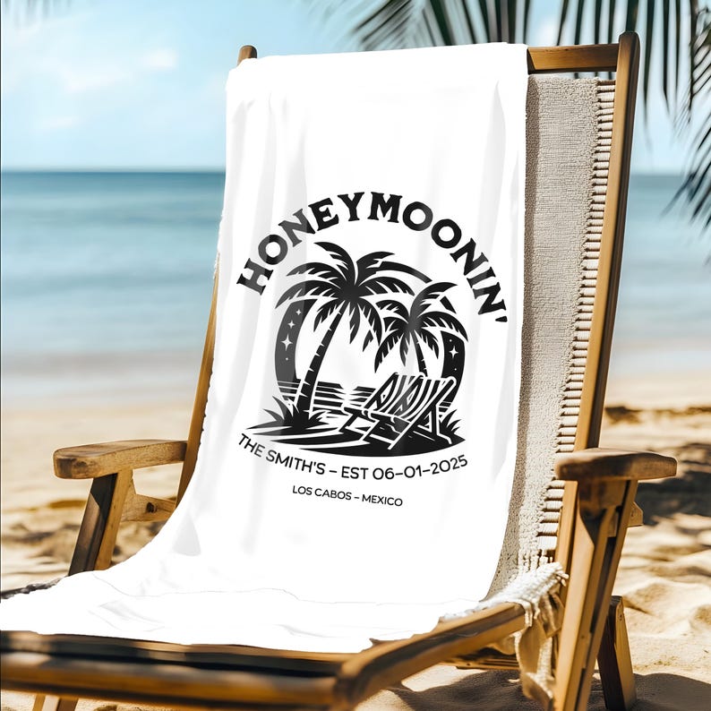 A white beach towel draped over a lounge chair on a sunny beach. The towel features a black tropical design with palm trees, two lounge chairs, and stars, along with the phrase “Honeymoonin’” in bold letters. Below the design, it is personalized with “The Smith’s – EST 06-01-2025” and “Los Cabos – Mexico.” The ocean and palm tree shadows create a peaceful, tropical setting.