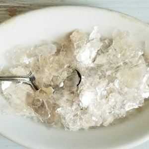 May include: A white oval dish filled with iridescent mica flakes. A silver spoon is resting on the edge of the dish.