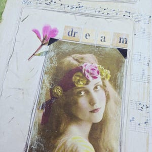 May include: Mixed media artwork with a vintage portrait of a woman wearing a floral headband. The word "dream" is spelled out in letter tiles above the portrait. A pink flower and sheet music fragments are included in the collage.