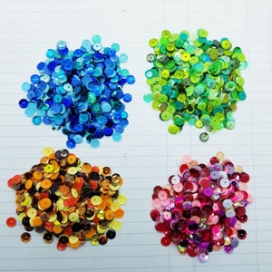 May include: Four piles of colourful round sequins. The piles are blue, green, orange, and red.
