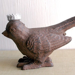 May include: A brown cast iron bird figurine with a silver crown on its head. The bird is standing on a light brown surface.