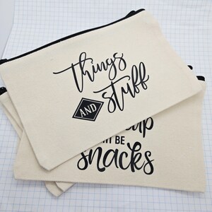 Peut inclure: Pochettes à fermeture éclair en toile écrue avec texte noir. La pochette supérieure porte l'inscription "things and stuff" en écriture cursive. La pochette inférieure porte l'inscription "snacks". Les pochettes ont des fermetures éclair noires.