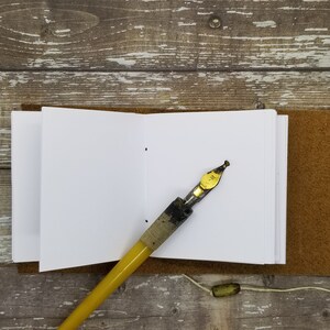 May include: A small, white, blank notebook with a brown leather cover and a yellow and black calligraphy pen. The notebook is open to a blank page.