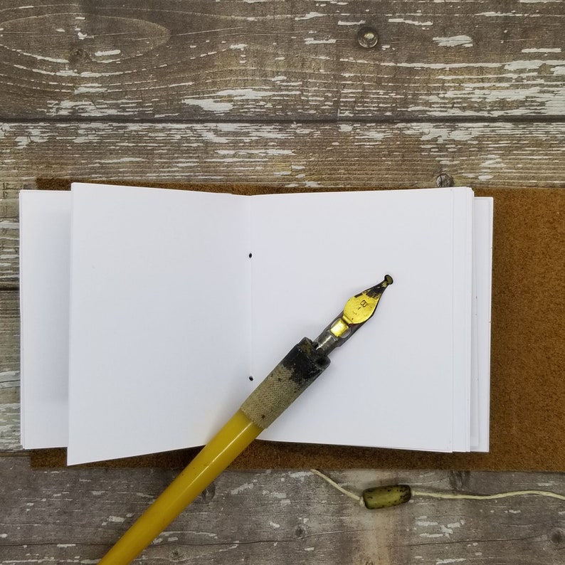 May include: A small, white, blank notebook with a brown leather cover and a yellow and black calligraphy pen. The notebook is open to a blank page.