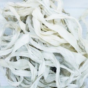 May include: A collection of ivory-coloured silk ribbon, with a slightly textured appearance. The ribbons are loosely arranged, creating a layered effect. The edges of the ribbons have a frayed texture. The overall aesthetic is soft and delicate.