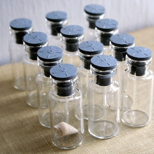 May include: A group of small, clear glass vials with grey caps and black rubber stoppers. The vials are arranged closely together, possibly for medical or scientific use. One vial contains a small, light-coloured object.