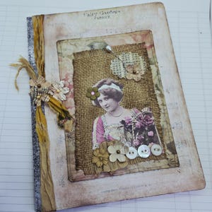 May include: A vintage-style handmade journal. The cover is a collage with a sepia-toned image of a woman, burlap, fabric flowers, and buttons. The journal has a decorative spine and ribbon binding.