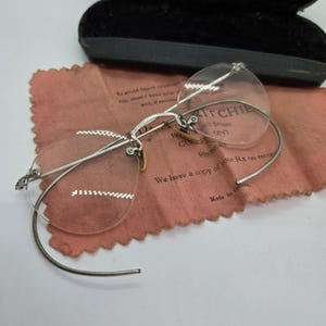May include: Antique rimless spectacles with a silver metal frame and clear lenses. The glasses are on a pink cloth with text, next to a black case. The lenses have a distinctive shape.