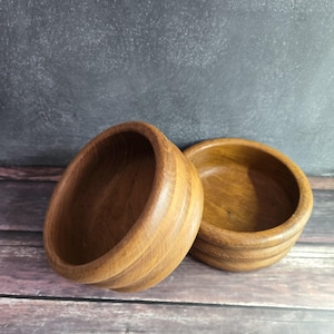 May include: Two wooden bowls with a natural wood grain finish. One bowl is tilted, revealing its interior, while the other rests upright. The bowls have a simple, classic design, suitable for serving or display.