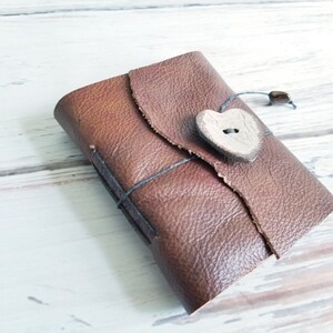 May include: A brown leather journal with a heart-shaped button closure. The journal is tied with a black string and has a rustic, handmade look.