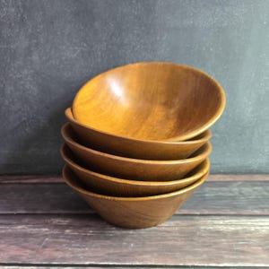 May include: Five small, wooden bowls stacked together. The bowls are a warm, golden brown colour, showing the wood grain. They are stacked on top of each other, with the top bowl slightly angled. The background is a dark grey surface.