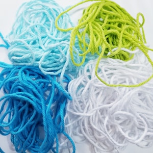 May include: Four skeins of yarn in different colours: light blue, blue, white, and green. The yarn is arranged in a pile.