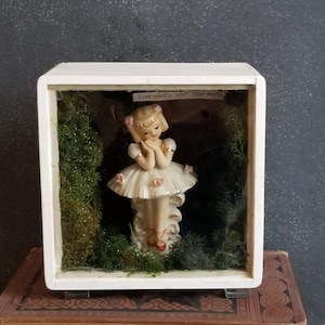 May include: A white shadow box containing a porcelain figurine of a ballerina wearing a white tutu and red shoes. The box is filled with green moss and the text "live gently upon this earth" is visible on a piece of paper inside the box.