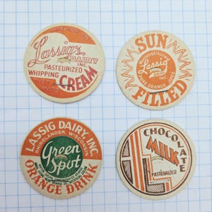 May include: Four vintage Lassig's Dairy milk bottle caps. The caps feature text and graphics in orange, green, and brown. The caps advertise whipping cream, orange juice, Green Spot orange drink, and chocolate milk.