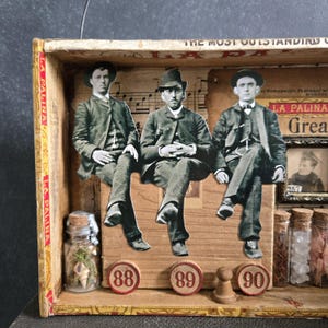 May include: A vintage wooden box art piece with three seated figures in monochrome. The box contains small glass jars with corks, and the numbers 88, 89, and 90. The text "LA PALINA" and "THE MOST OUTSTANDING" are visible.