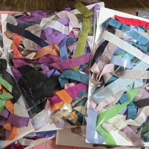 May include: A selection of colourful ribbon offcuts in clear plastic packaging. Ribbons in shades of purple, pink, orange, blue, green, and black. The ribbons vary in texture and width, ideal for crafts and embellishments.