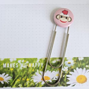 May include: A large silver paper clip with a pink button featuring a sock monkey face. The paper clip is attached to a page with the words "MAKES ME HAPPY" printed on it, with a background of daisies.