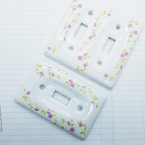 Peut inclure: Deux interrupteurs de lumière en céramique blanche avec un motif floral. Le motif présente de petites fleurs violettes et des vignes vertes.