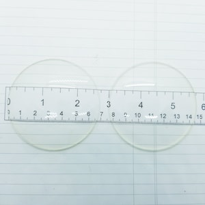 May include: Two clear glass circles, each measuring approximately 5 cm in diameter, are placed on a white surface with a ruler underneath. The ruler is marked with numbers from 0 to 6.