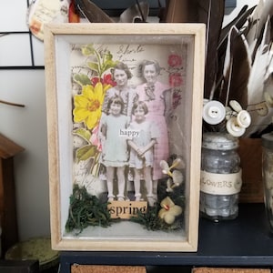 May include: A vintage photograph of three girls with flowers and the word "happy" in a shadow box with a wooden frame. The shadow box also includes green moss and the word "spring" in gold letters.