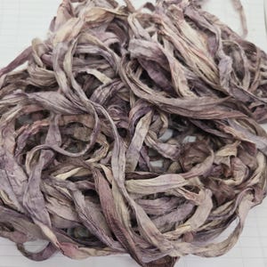May include: A tangled pile of hand-dyed silk ribbon featuring a variegated colour palette of muted mauve, dusty purple, and soft cream tones. The fabric strips have a crinkled, textured appearance with raw, frayed edges throughout the entire bundle.