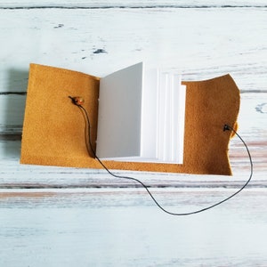 May include: A small, brown leather-bound journal with a white paper interior and a black cord closure. The journal is open to reveal the blank pages inside.