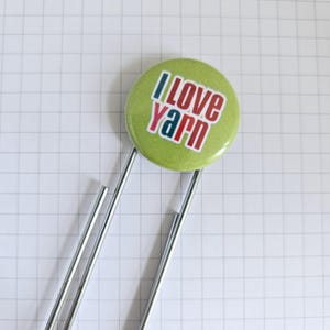 May include: A green button with the text "I LOVE YARN" in red, blue, and white. The button is attached to a silver paper clip, set against a white grid background. A craft-themed accessory.