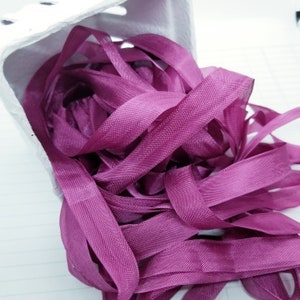 May include: A pile of deep purple satin ribbon, with a soft sheen, is spilling out of a white container.